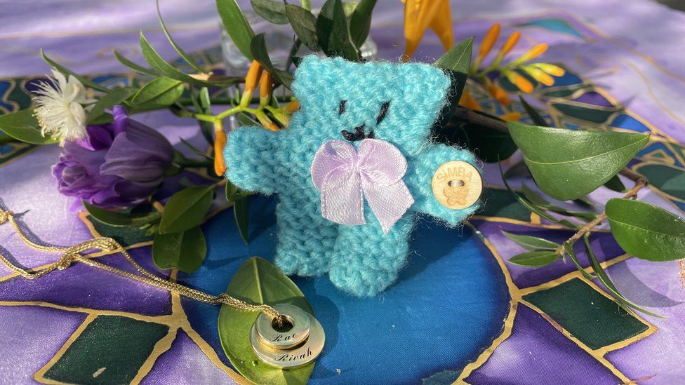 Knitted blue teddy bear and flowers alongside a necklace engraved with the names Rivah and Rae.