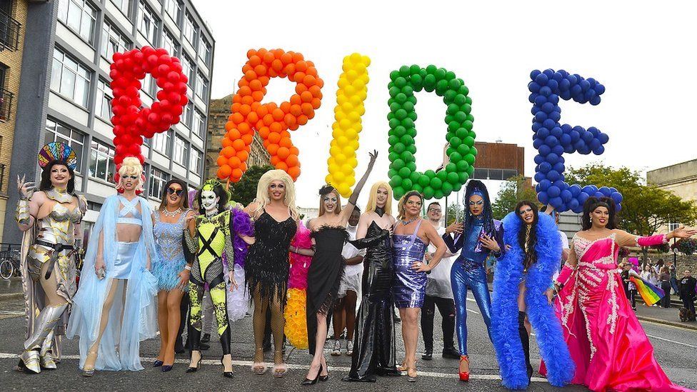 A group of participants stand in front of red, orange, yellow, green and blue balloons which spell out "Pride"