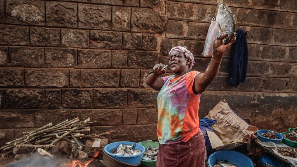 Kenyan woman posing with a fish. She is holding up the fish in one hand and a knife in the other. There is a brick wall behind her and tubs of fish.