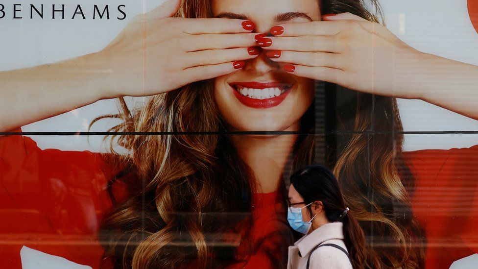 Woman with surgical mask walks past a large Debenhams window poster