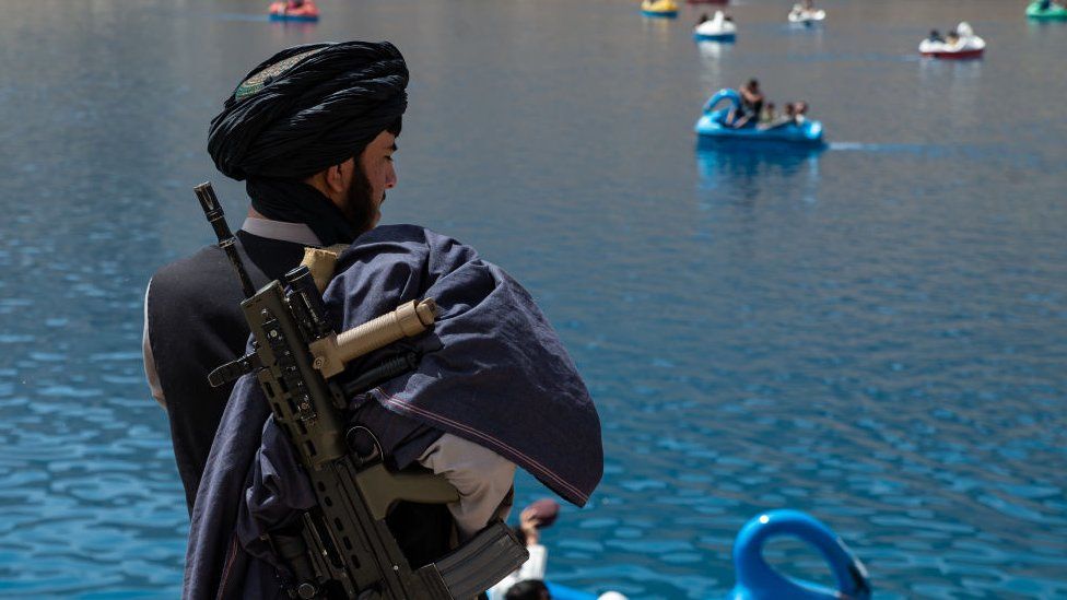 A guard with a gun on his back stands near a lake where children and adults play in Band-E-Amir national park