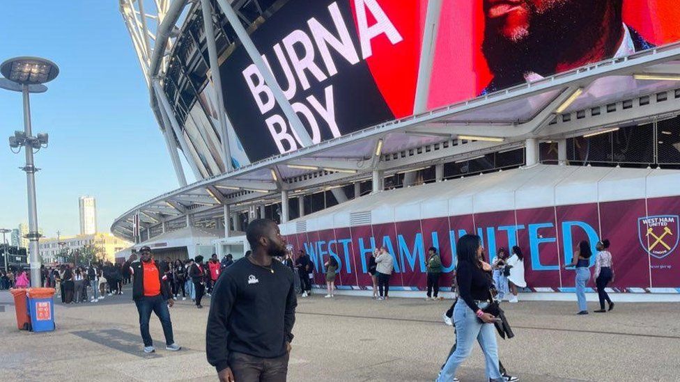 Kojo Amankwah wearing a black jumper stood in front of the London Stadium looking up at a sign reading "Burna Boy" at the top of the stadium