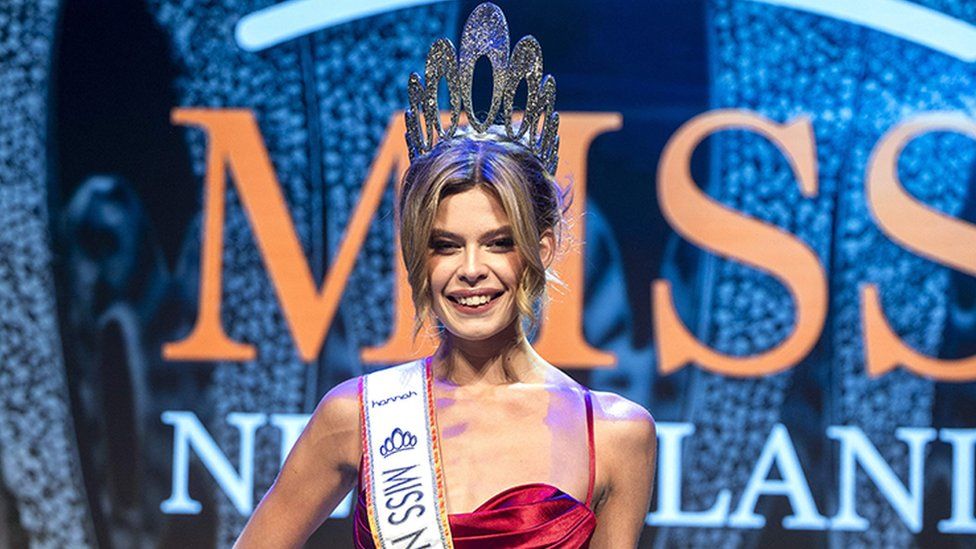 Rikkie Valerie Kolle after being crowned Miss Netherlands on stage. She is wearing a red dress and has a white sash around here around here saying Miss Netherlands 2023. She has a big silver tiara on her head and flowers in her left hand. The background has a sign in orange and white saying Miss Netherland