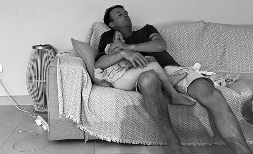 El marido de Hollie, Mike, acurrucando a Sydney en el sofá. La foto tiene un tratamiento en blanco y negro.