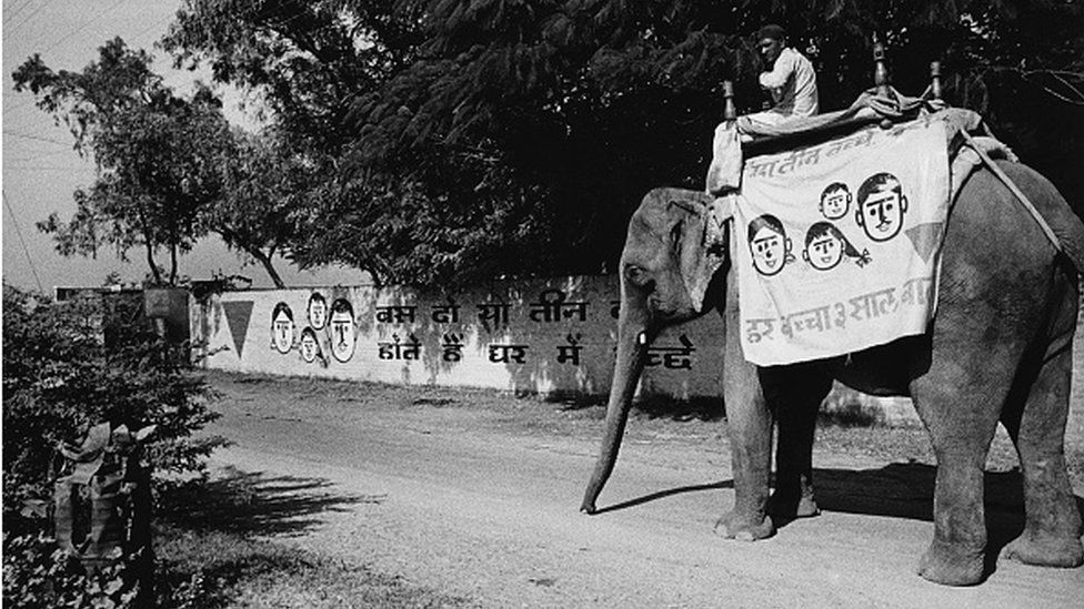 An elephant bearing the red triangle symbol of the Lal Tikon Fund to publicise birth control and family planning, enters a village to spread the news and offer information.
