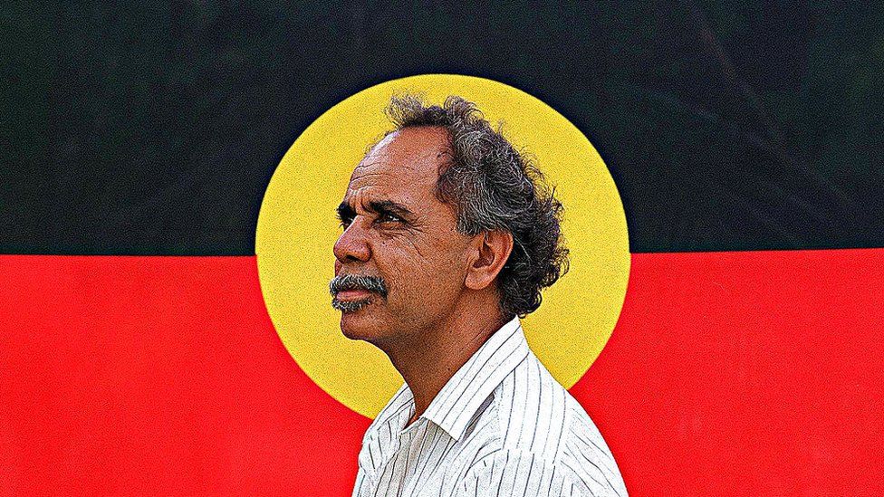 Side view of Harold Thomas in 1994 framed in the centre of an Aboriginal flag backdrop