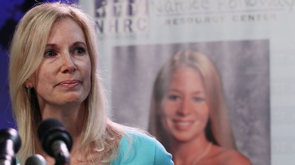 Beth Holloway with blonde hair and green shirt in front of microphones, photo of Natalie Holloway behind her