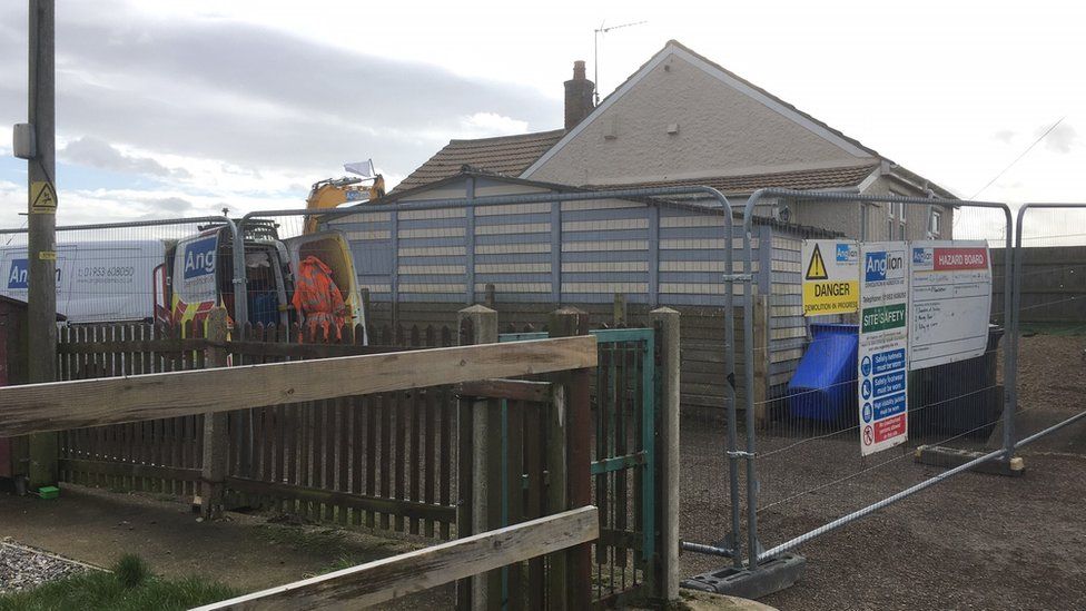 Single-storey chalet with demolition crews outside and metal fencing