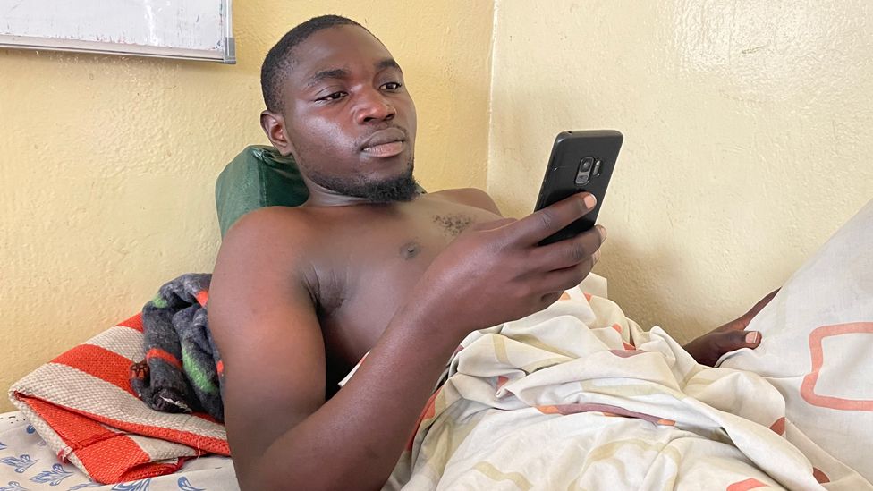 Mundeke Kandundao lies in a hospital bed, shirtless, looking at his mobile phone