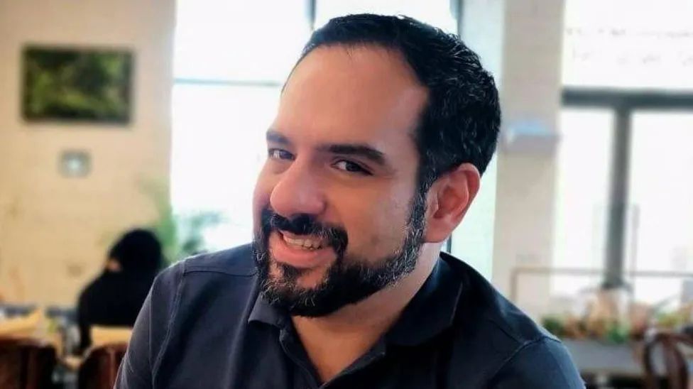 A man with black hair and a black beard smiles at the camera, he's wearing a blue polo shirt and appears to be in a cafe.