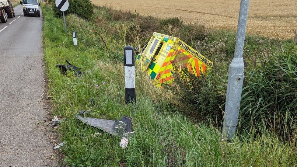 An ambulance at the bottom of a roadside ditch, leaning towards its right