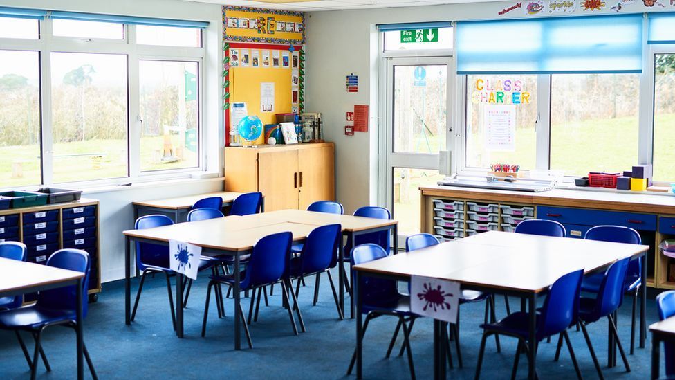 An empty school classroom, there are three tables visible with blue chairs tucked underneath. In the background drawers and cabinets line the walls and a grassy field is visible through windows.