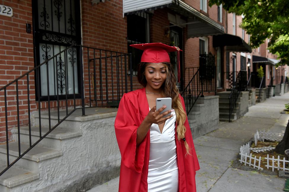 18-летняя Франсина Таунс, одетая по случаю окончания средней школы Вудлона, смотрит в свой мобильный телефон, стоя перед своим домом