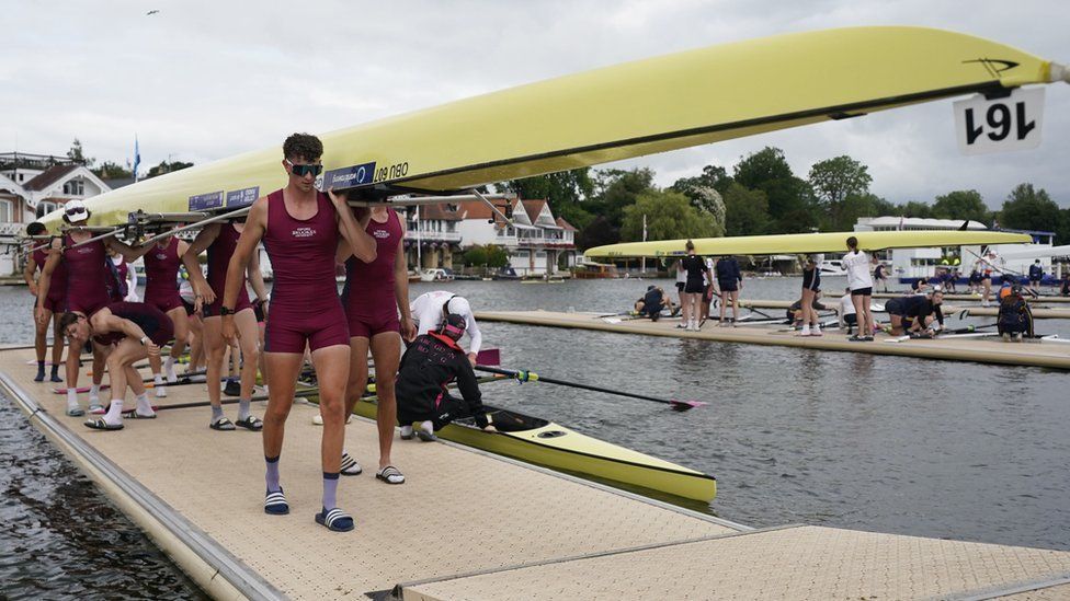 Rowing crew in burgundy race suits carry a pale yellow rowing boat off a pontoon