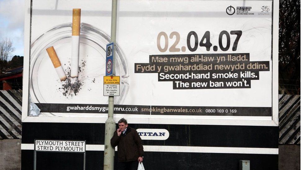 A man stands in front of an advert about banning smoking, smoking a cig