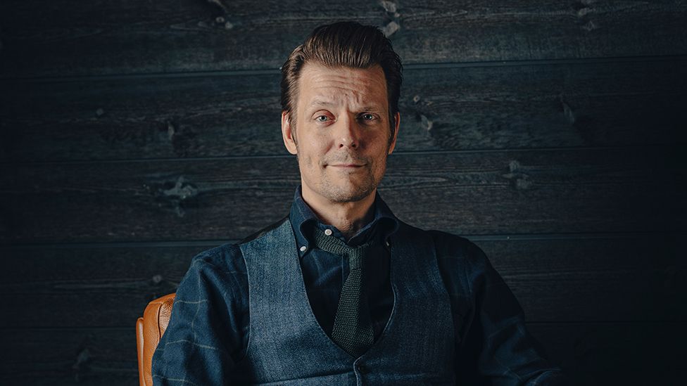 A smartly-dressed man in a navy blue shirt and matching waistcoat from a three piece suit sits in front of a dark brick wall. He has hair that is very neatly cut and styled into a short, rockabilly-ish pompadour style. He's got one eyebrow raised and looks quizzical.