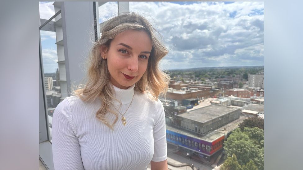 Zsofia Vakos, standing in a building looking over the shopping centre. She has blonde hair, red lipstick, a white polar neck and a gold necklace 