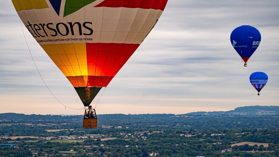 Three hot air balloons high in the air above the city. One of them is colourful and lit up with the glow of the fire used to ignite the fuel