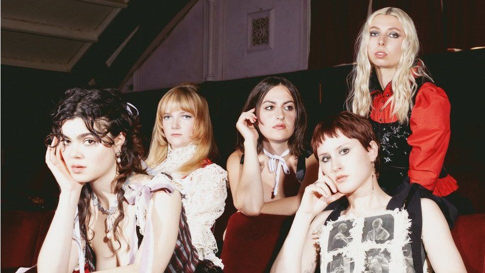 The Last Dinner Party (L-R): Abigail Morris, Emily Roberts, Aurora Nishevci, Lizzie Mayland and Georgia Davies. The five young women are pictured inside wearing outfits inspired by the past - lacey tops, corsets and neck ties. Abigal has curly brown hair tied back and leans forward towards the camera, her face resting on her right hand. Behind her is Emily, who has long strawberry blonde hair worn loose. To her left is Aurora, who has shoulder length brown hair and looks away from the camera, her right hand touching her face. In front of her is Lizzie who has cropped reddish brown hair and brown eyes. Georgia sits at the back, taller than everyone else, with long platinum hair and metallic eyeshadow.