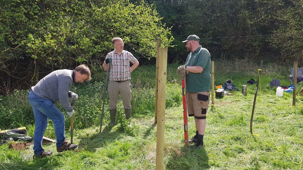 Three men standing in a field using tools to build a shelter from wood
