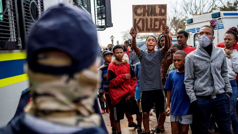 A resident holds a placard that reads: "Child Killer" next to members of the South African Police Service (SAPS) outside the SAPS offices in Eldorado Park, near Johannesburg, on August 27, 2020, during a protest by community members after a 16-year old boy was reported dead
