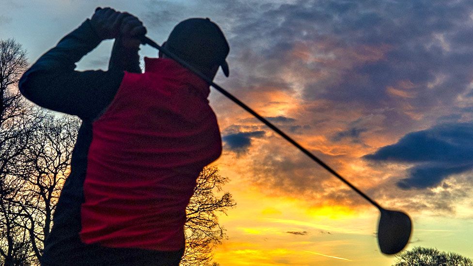 A golfer hits a ball on a golf course as the sun rises