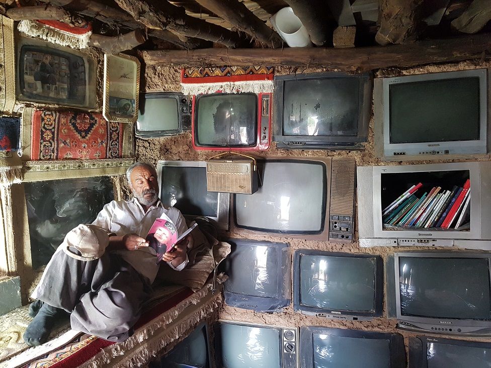 A man reading in his home next to a wall embedded with televisions