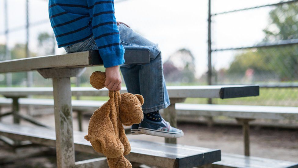 A young boy sitting by himself on a stand of benches, holding a teddy bear