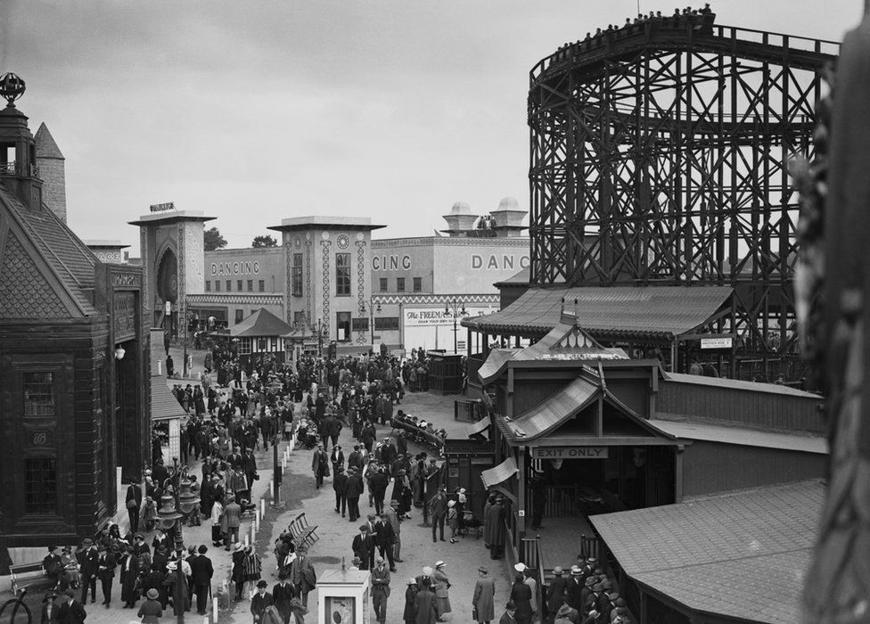 Black and white image of people walking through the amusement park with dancing hall and roller coaster in the background