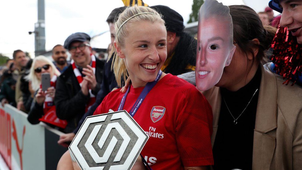 Leah laughing with a fan wearing a mask of her face after an Arsenal game
