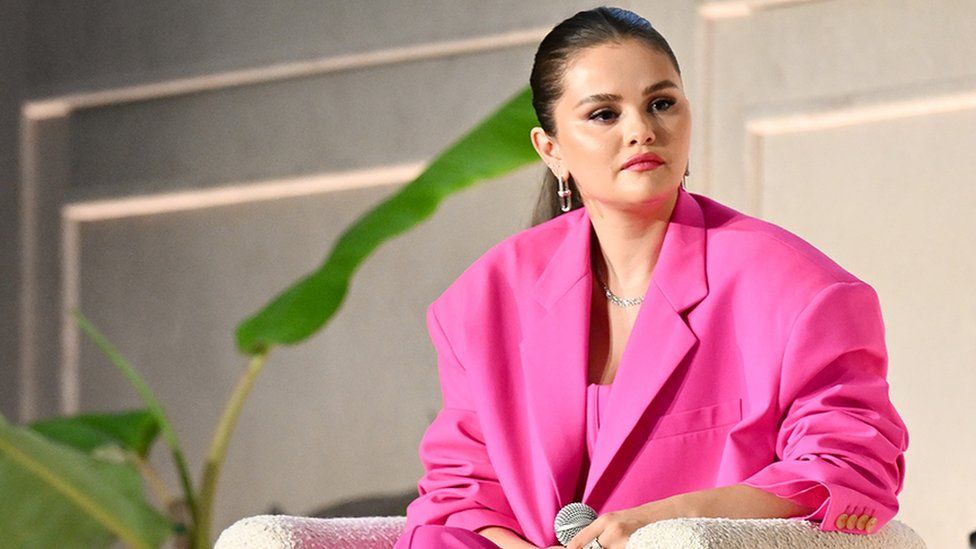 Selena Gomez at the Music + Health Summit presented by Universal Music Group and Thrive Global at 1 Hotel on September 19, 2023 in West Hollywood, California. Selena, a woman in her early 30s, has her long dark hair tied back in a slick pony tail. She wears a bright pink oversized suit with dangly gold earrings and a silver chain necklace. She wears pink lipstick and black eyeliner and is sitting in a cream chair, looking over her left shoulder towards someone out of shot. A potted banana plant is next to her seat and she holds a microphone in her left hand in her lap.