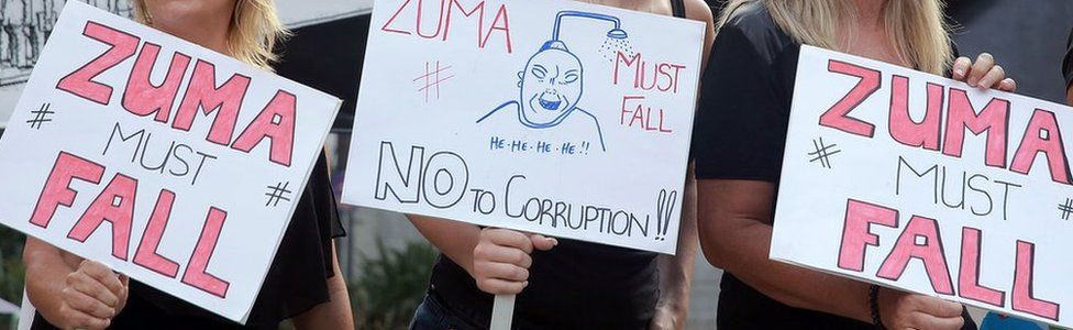 Three women holding placards at a protest calling for South Africa's president to step down.