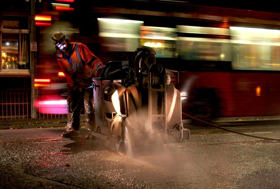 A construction worker works through the night as a red bus passes him by