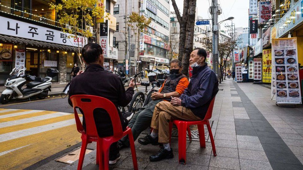 Three men chat as they sit on chairs on a pavement in Seoul on November 4, 2021