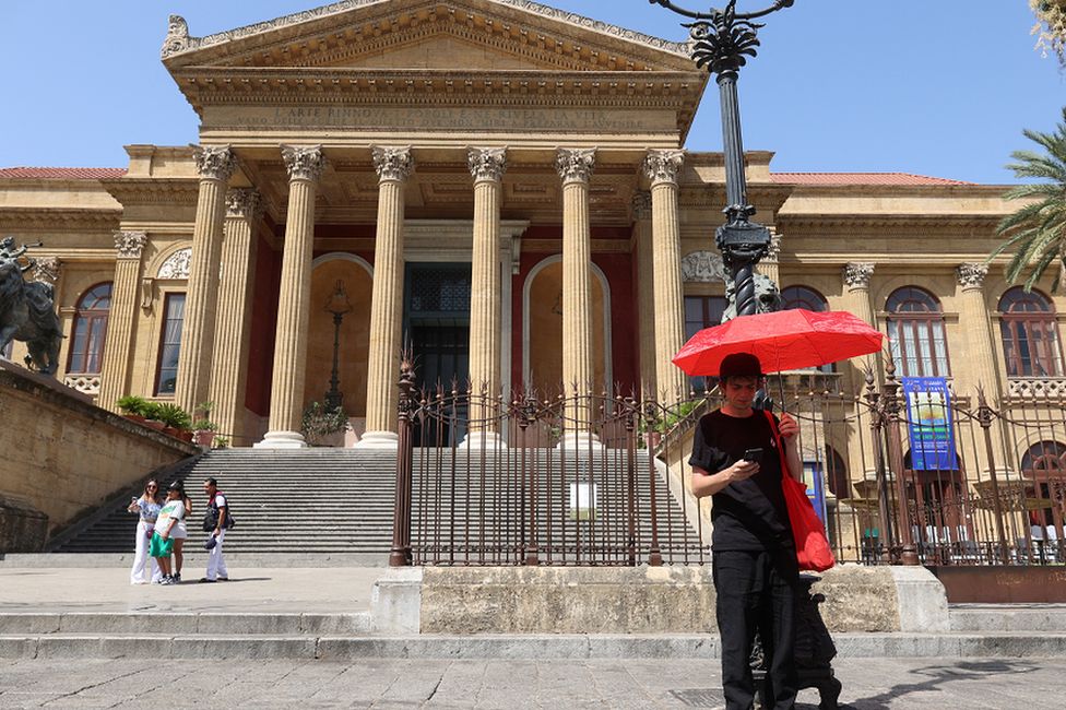 Мужчина стоит с красным зонтом на площади Верди перед театром Массо Витторио Эмануэле в Палермо, вторник