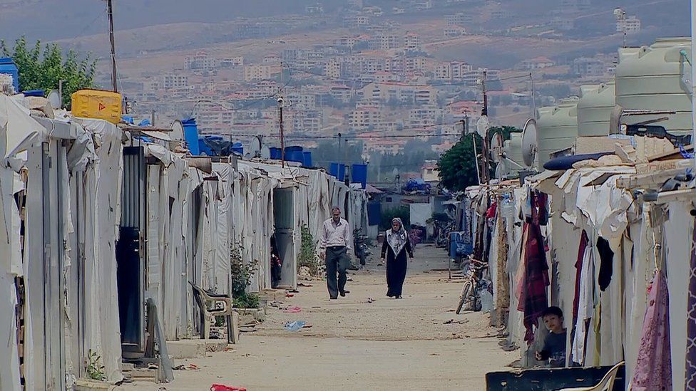 Two rows of tents in a refugee camp for Syrians living in Lebanon. A man and a woman walk along a path between the two rows of tents.
