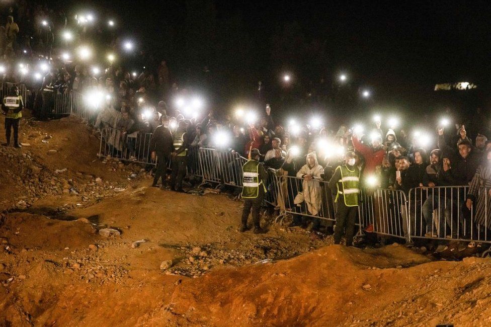 People stand behind a cordon. They use their mobile phones as torches. February