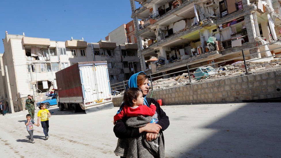 woman carrying child walks past destroyed building but another one in the background is much less damaged