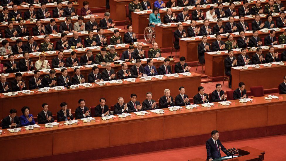 Delegates applaud a speech by President Xi Jinping at China's Communist Party Congress in Beijing. Most are men in dark suits, with a handful of women dotted around.