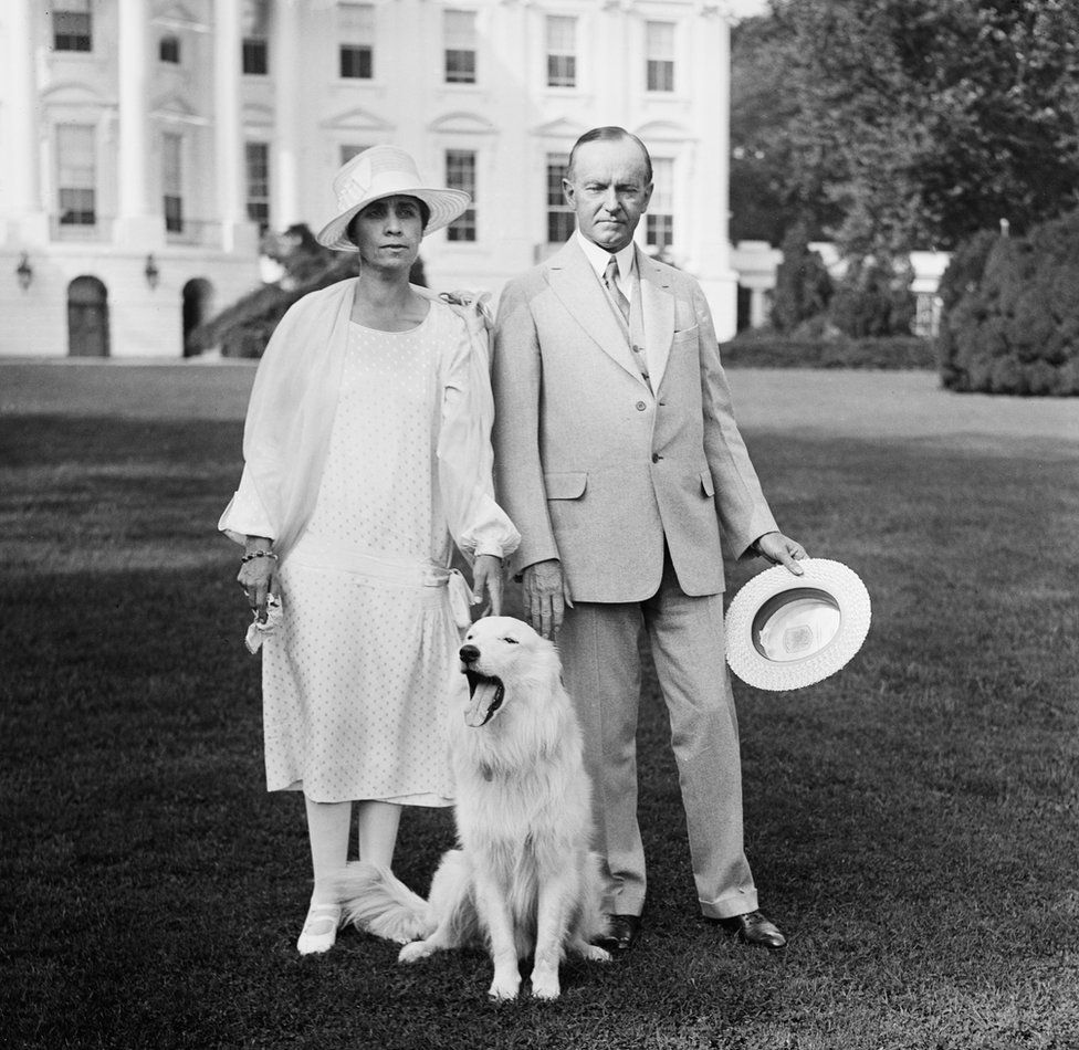President Calvin Coolidge and First Lady Grace Coolidge stand in front of the White House with a dog in front of them that is yawning