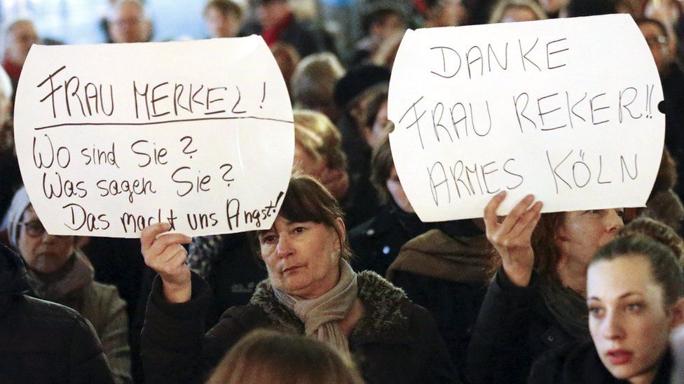 A woman holds a placard saying "Mrs Merkel: Where are you? What do you say? This alarms us" while another's reads: "Thanks Mrs Reker; poor Cologne"