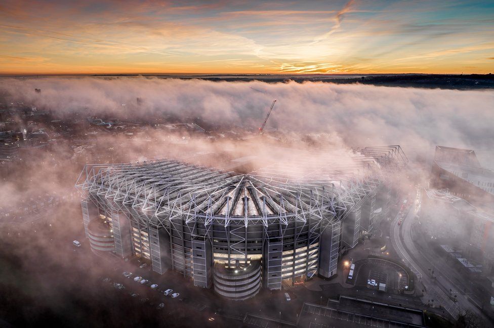 A football stadium emerges from low cloud with orange clouds and blue skies visible beyond