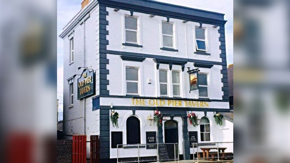 Exterior of The Old Pier Tavern in Burnham-On-Sea. It is a white building with a blue paint finish and two pub signs hanging from the walls