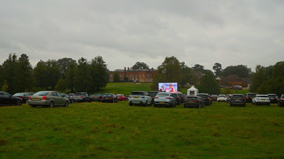 Cars gathered in front of a large screen showing a wedding.