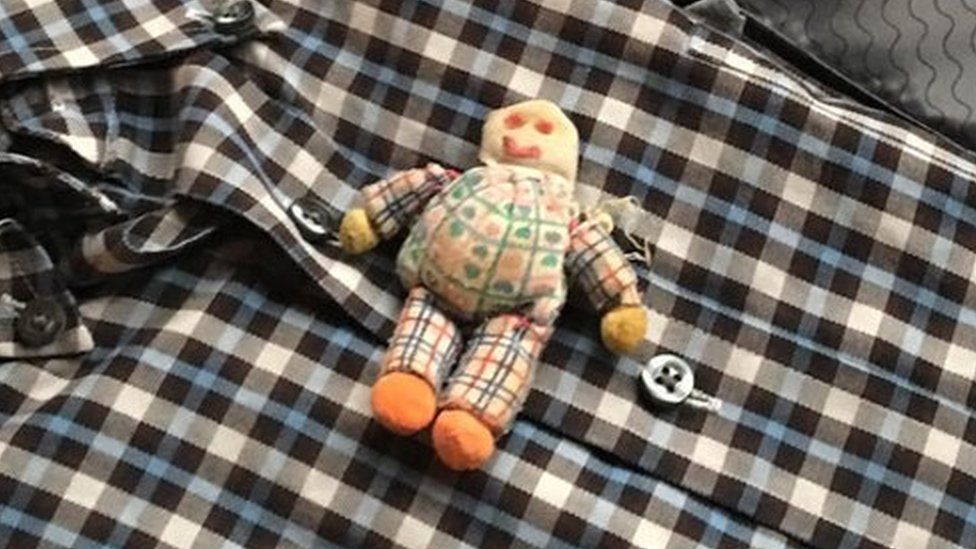 An image of a doll packed in a suitcase on top of a shirt.
