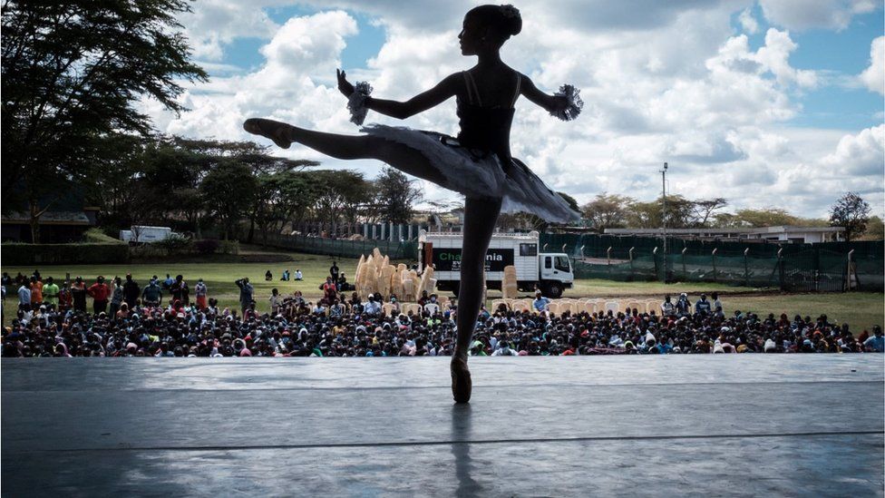 A ballet dancer standing on pointe in front of a crowd of people outside. The skies are cloudy with some blue creeping through.