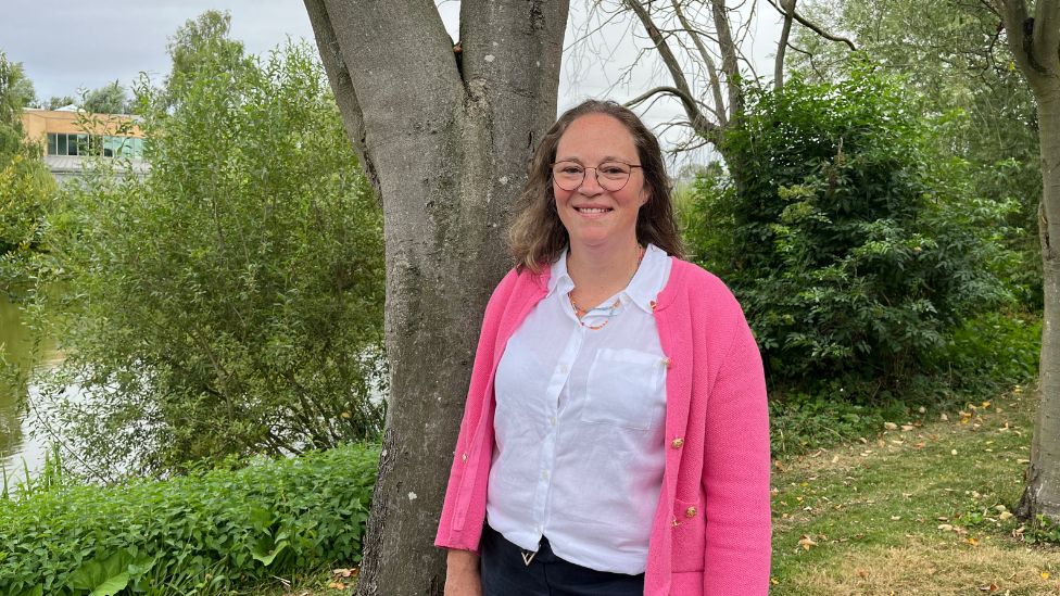 Vhari Russell pictured outside on the grass, stood in front of a tree trunk with a lake in the background. She wears thin framed circle glasses, has shoulder length brown hair and and smiles at the camera. She is wearing a white blouse and a bright pink jacket with gold buttons.