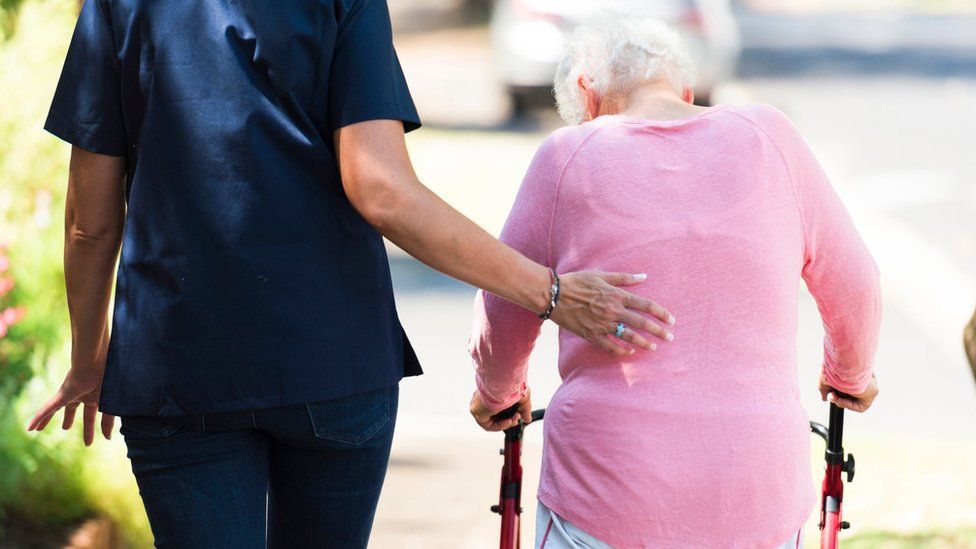 A caregiver helps an elderly woman walk with her walking aid outdoors
