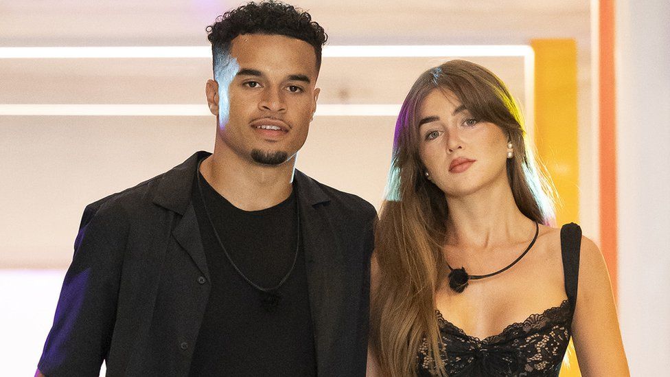 Toby Aromolaran and Georgia Steel pictured during Love Island Games. Toby is a mixed race man in his 20s with short dark hair with a goatee beard. He wears a dark blazer over a black top. Georgia is a white woman in her 20s with long brown hair. She wears a black bustier-style dress. They are pictured leaving the villa together