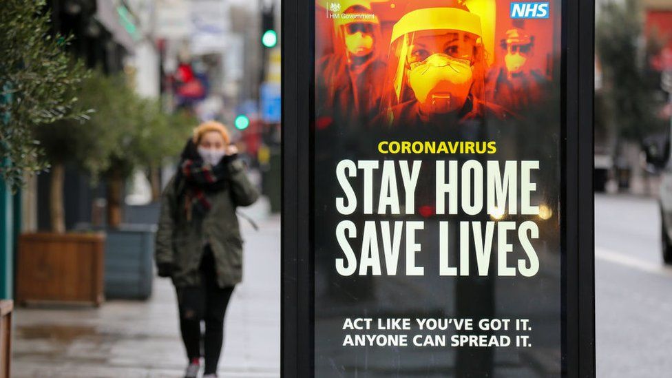 Woman walking past a Stay Home, Save Lives poster